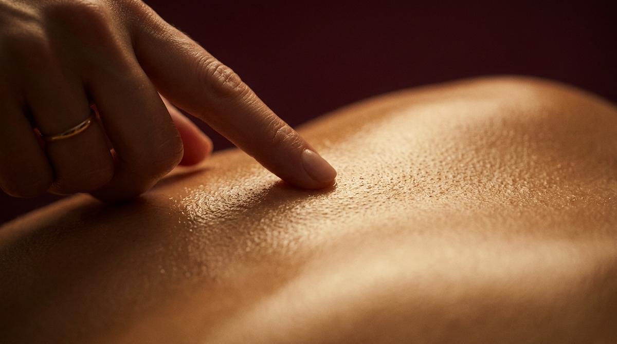 Plano macro de un dedo rozando suavemente una espalda masculina con aceite de masaje dorado.